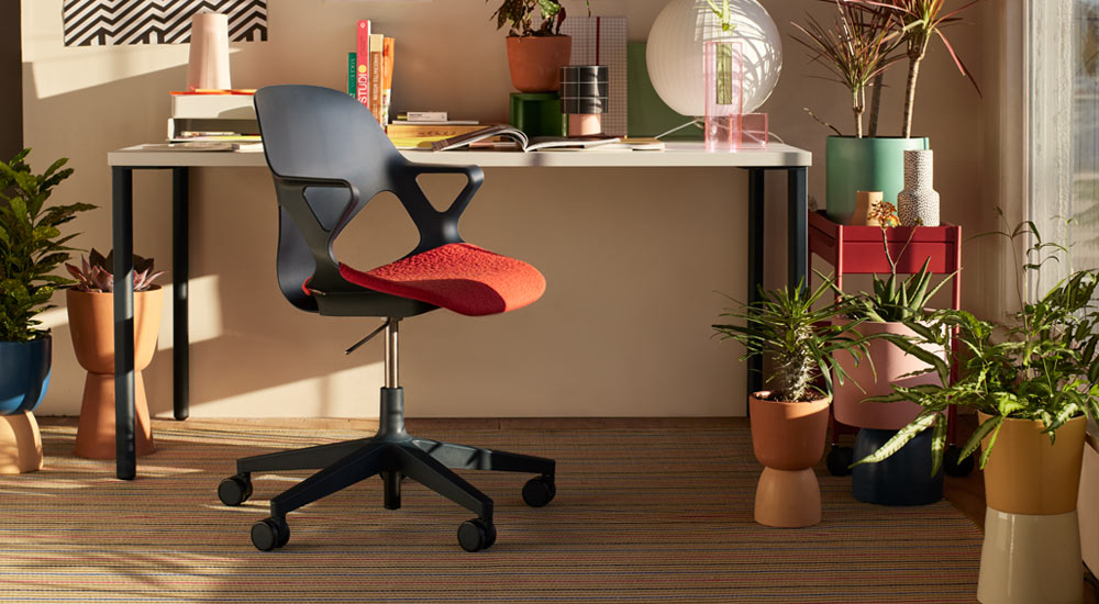 A black Zeph office chair with a red seat pad is positioned in front of a Herman Miller OE1 desk. Set in a home office environment, with a red OE1 trolley and a selection of indoor plants in colourful pots.