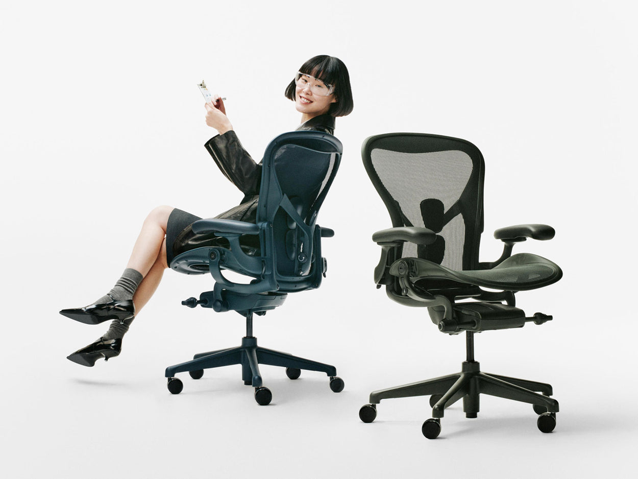 A woman sat on a Nighfall blue Herman Miller Aeron ergonomic office chair alongside another Aeron in Jasper green in a white studio.