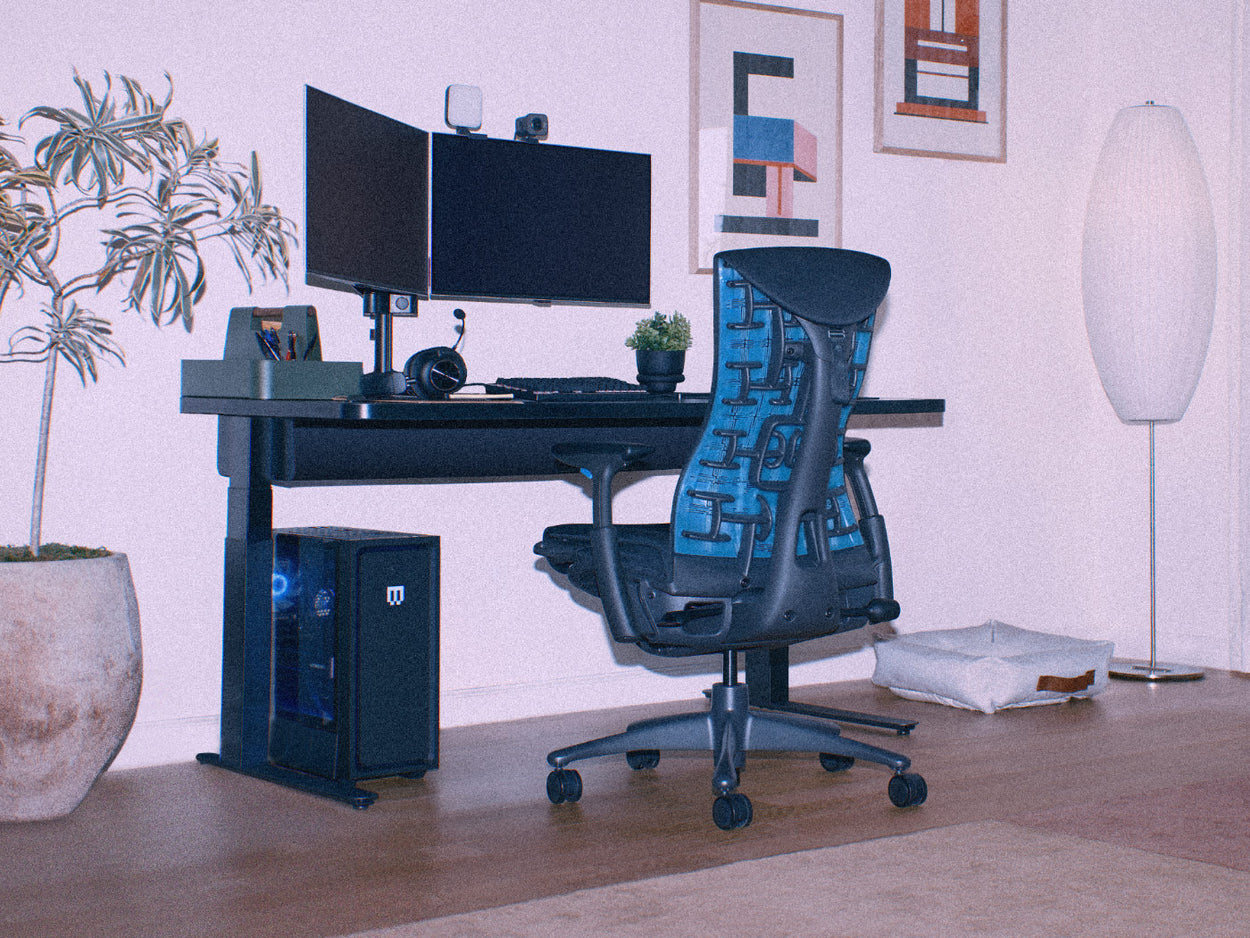 A grainy image of a black and cyan blue Herman Miller Embody gaming chair as part of a home PC gaming setup including a Nevi sit-stand gaming desk.