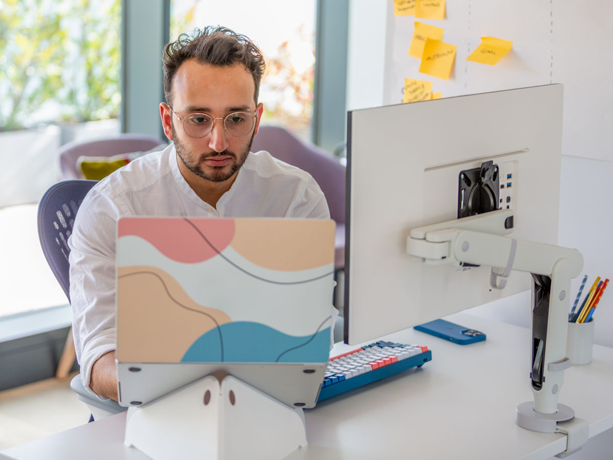 Photo of man sat at desk with monitor in front of him on Ollin monitor arm and laptop in Oripura stand, showing that your eyes should be directly in front of screen two thirds up the screen.