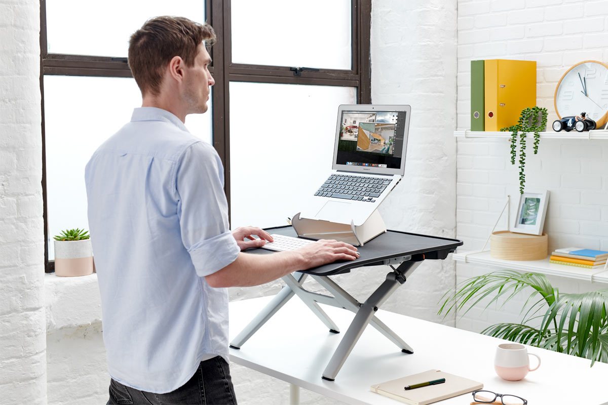 A man stood in front of a white desk with a Monto sit stand by Herman Miller positioned on top, holding a white Oripura laptop stand by Herman Miller.
