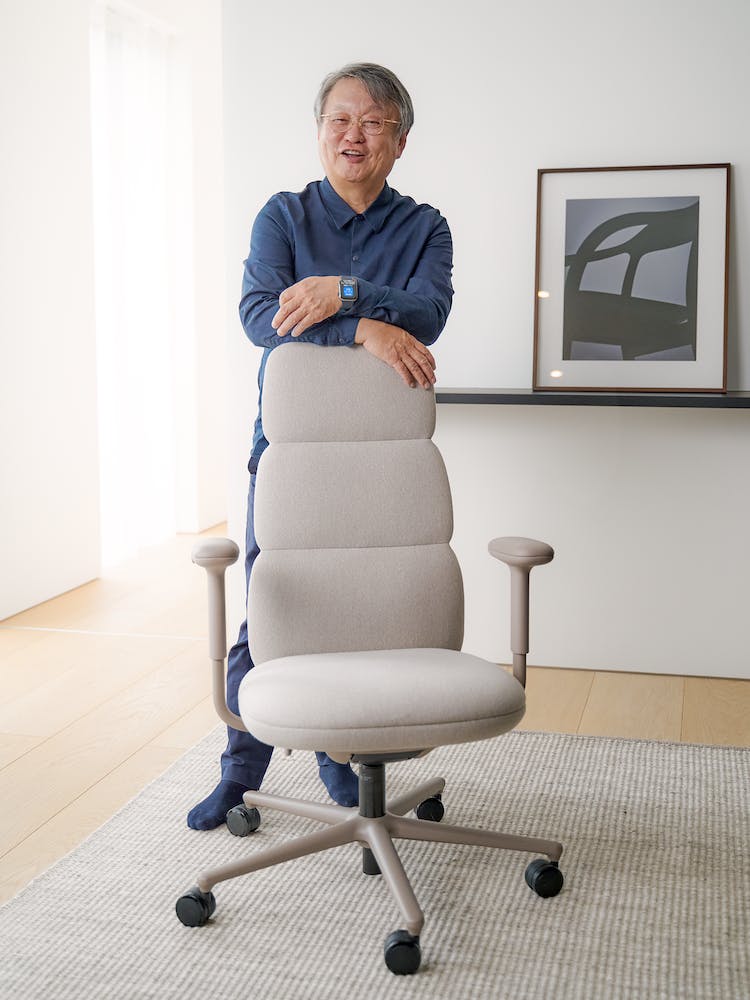 The designer, Naoto Fukasawa, leaning on a Herman Miller Asari office chair. The setting features a clean floor and wall in the background.