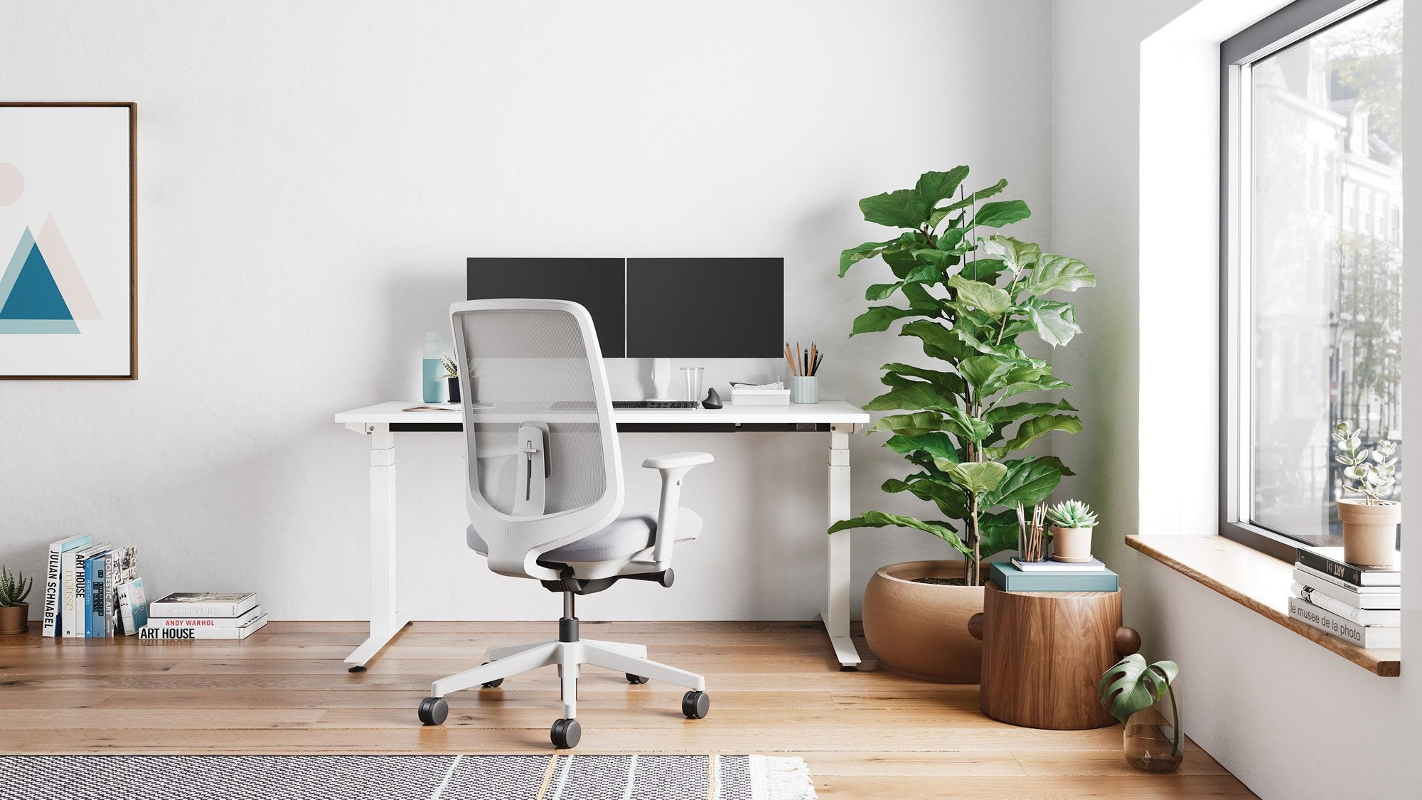 A grey Verus office chair by Herman Miller, positioned in front of a white Nevi standing desk by Herman Miller. Set in a home office environment, with wooden floors, white walls and indoor plants.
