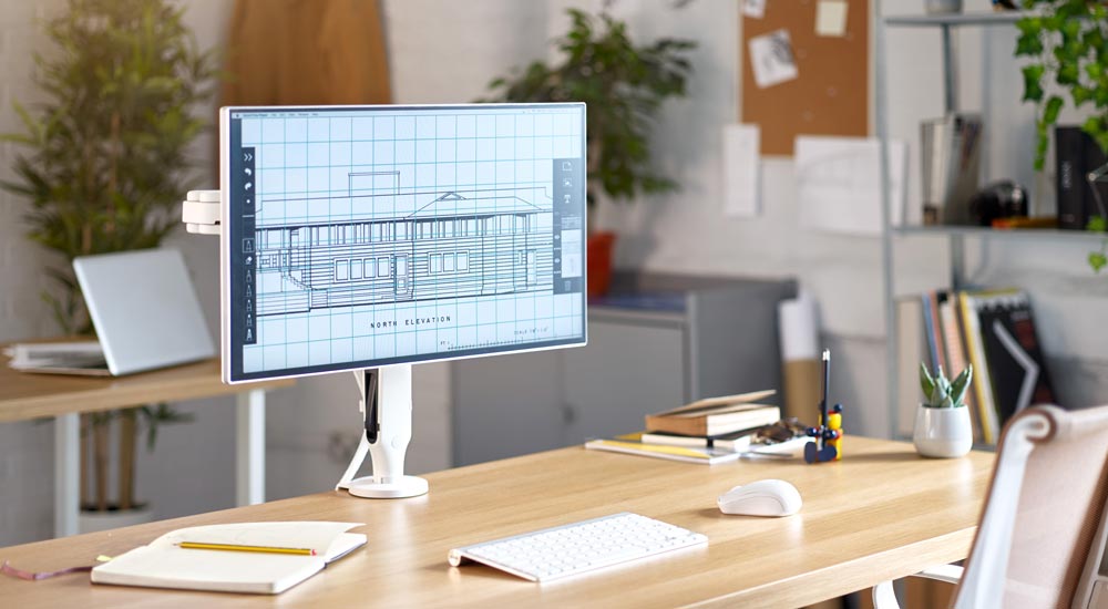 A white Ollin monitor arm by CBS, mounted on a wooden desk holding a computer screen, set in an office environment.