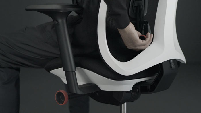 A video of a person sitting in a white and black Vantum gaming chair by Herman Miller, reaching behind the chair to adjust the back support.