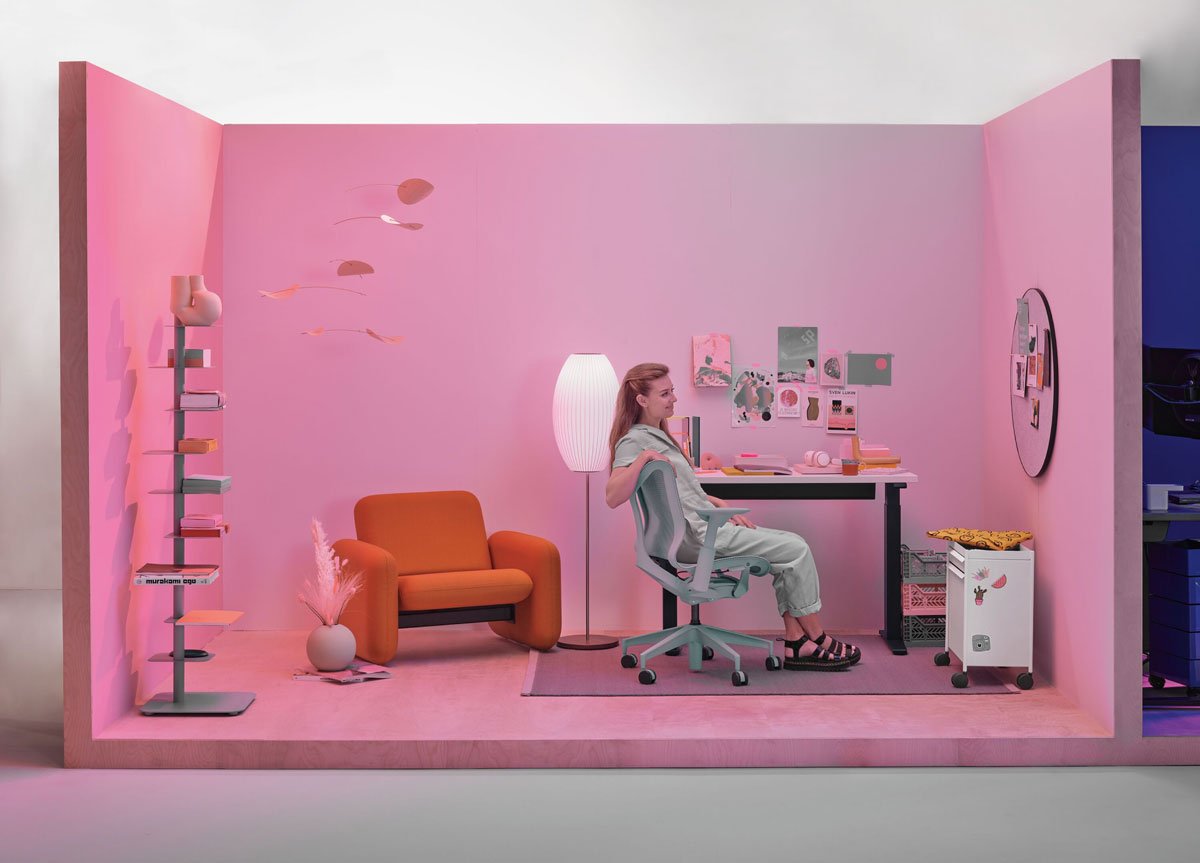 Home office setting with lady sat in Cosm low back office chair in glacier grey with compact sit stand desk with Wilkes arm chair in background