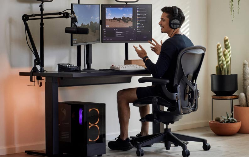 Gamer and streamer Xaryu in a gaming set up sat on Herman Miller Aeron gaming chair with black Nevi sit-stand gaming desk and dual monitors on Ollin moniter arms