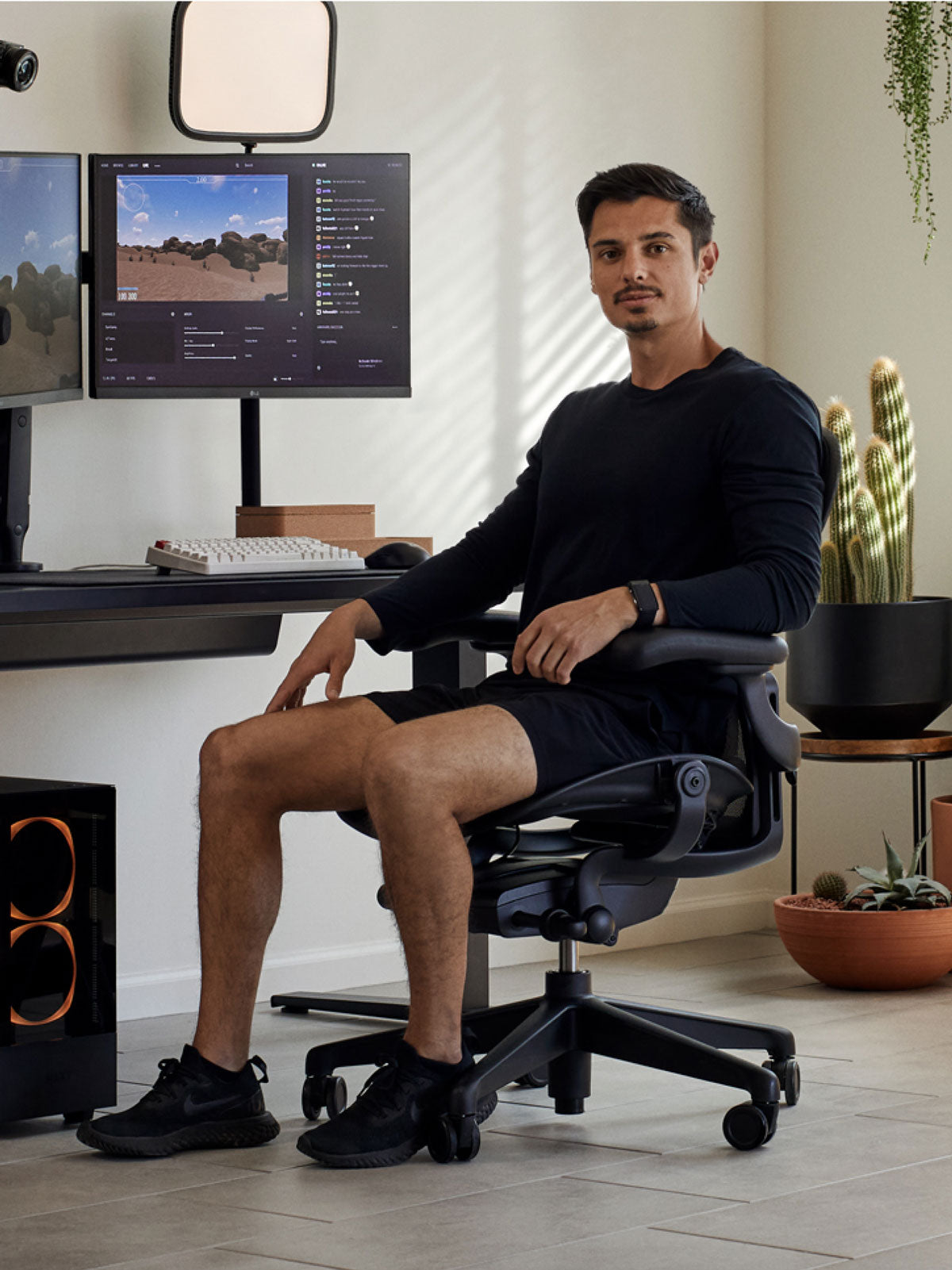 A man sitting in a black Aeron gaming chair by Herman Miller, positioned behind a black Nevi desk with a black Ollin monitor arm and screen. Set in an indoor space with tiled floors, white walls and indoor plants in terracotta pots.