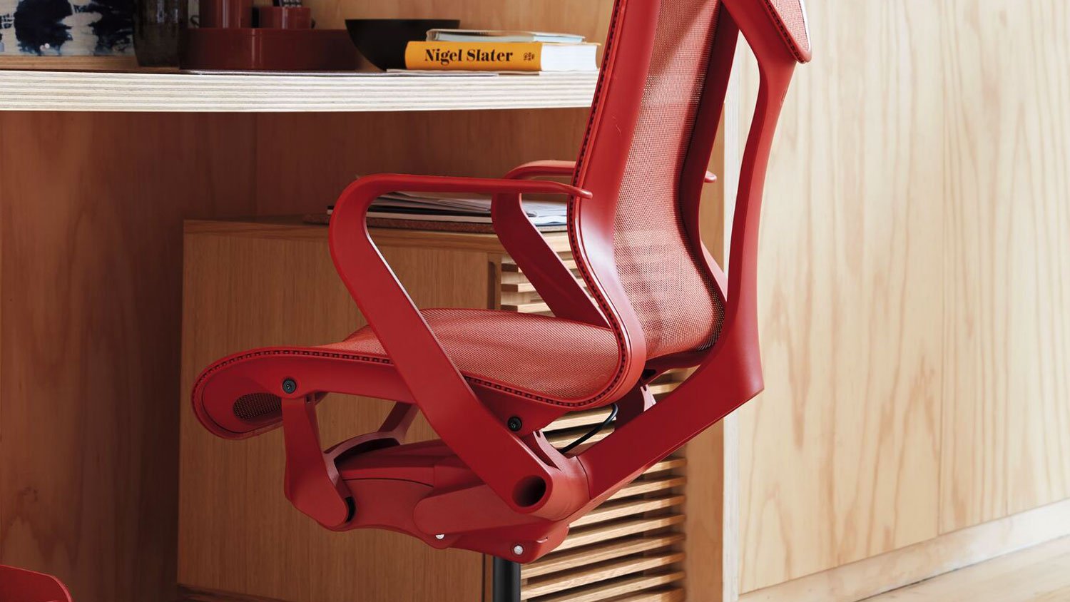 A red Cosm office chair by Herman Miller, is positioned under a desk. It is set in an indoor environment with hardwood flooring and walls, with a focus on the vibrant colour of the chair amidst the surrounding furniture.