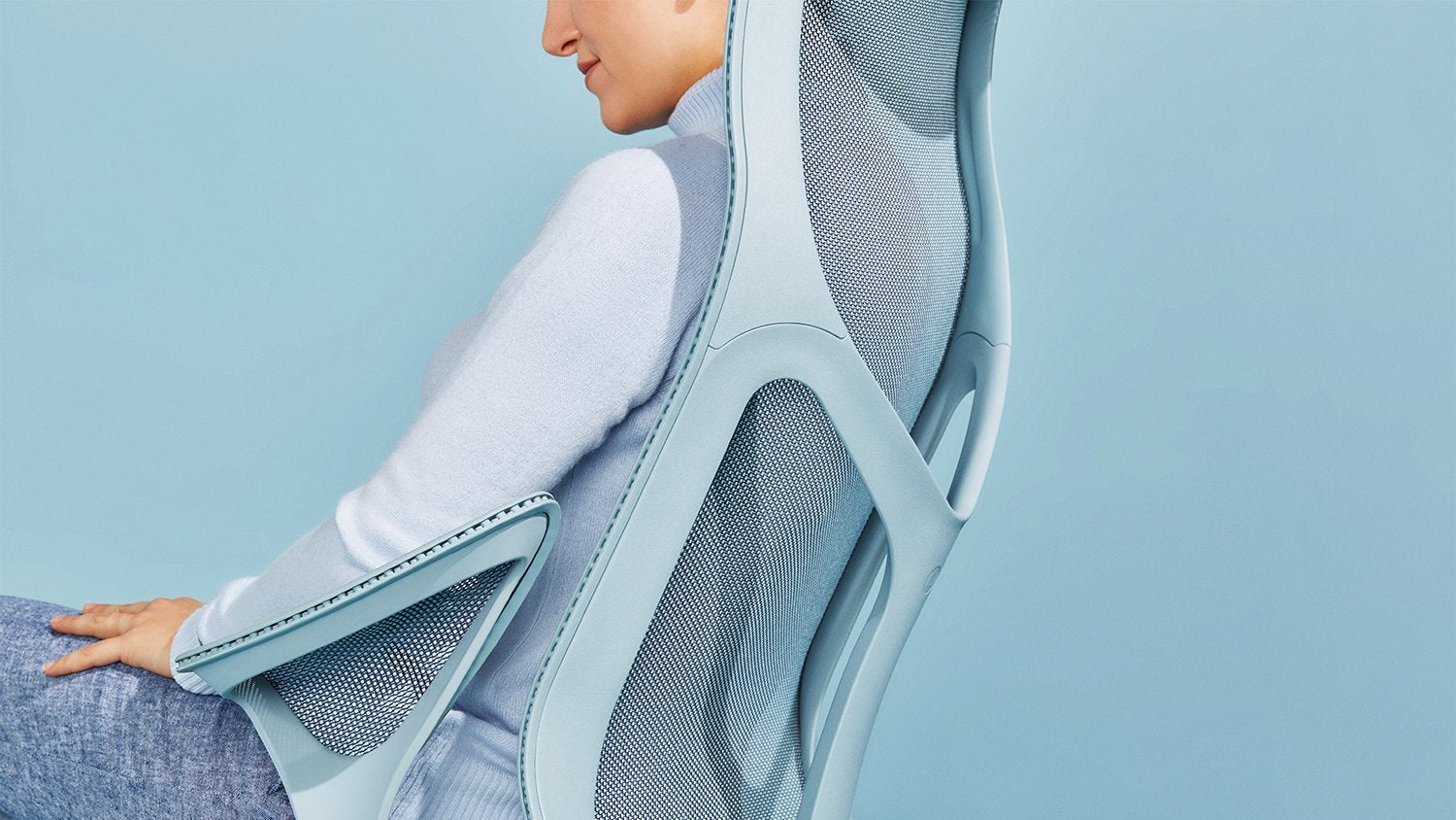 A person sitting in a mineral blue Cosm office chair by Herman Miller, dressed in a blue jumper and jeans in front of a plain pale blue background.