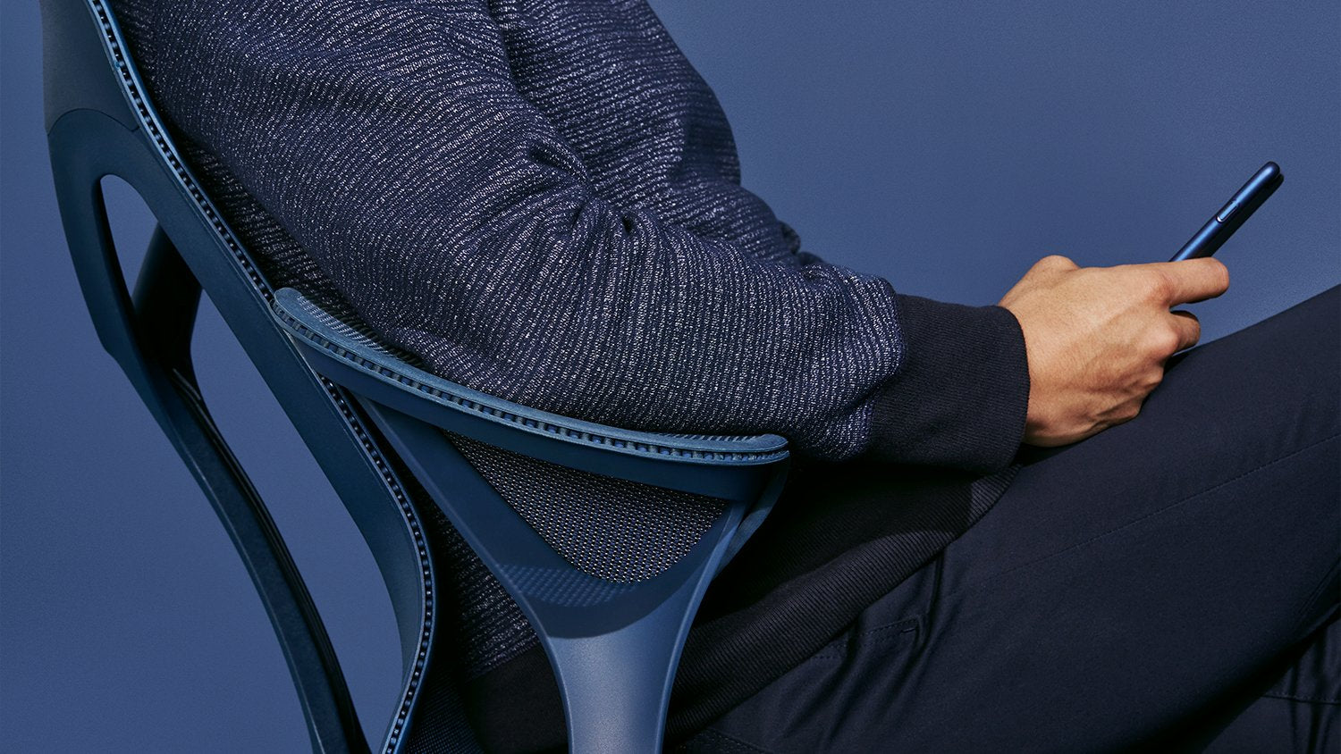 A person sitting in a nightfall blue Cosm office chair by Herman Miller. They are holding a phone and dressed in dark blue clothing in front of a plain dark blue background.