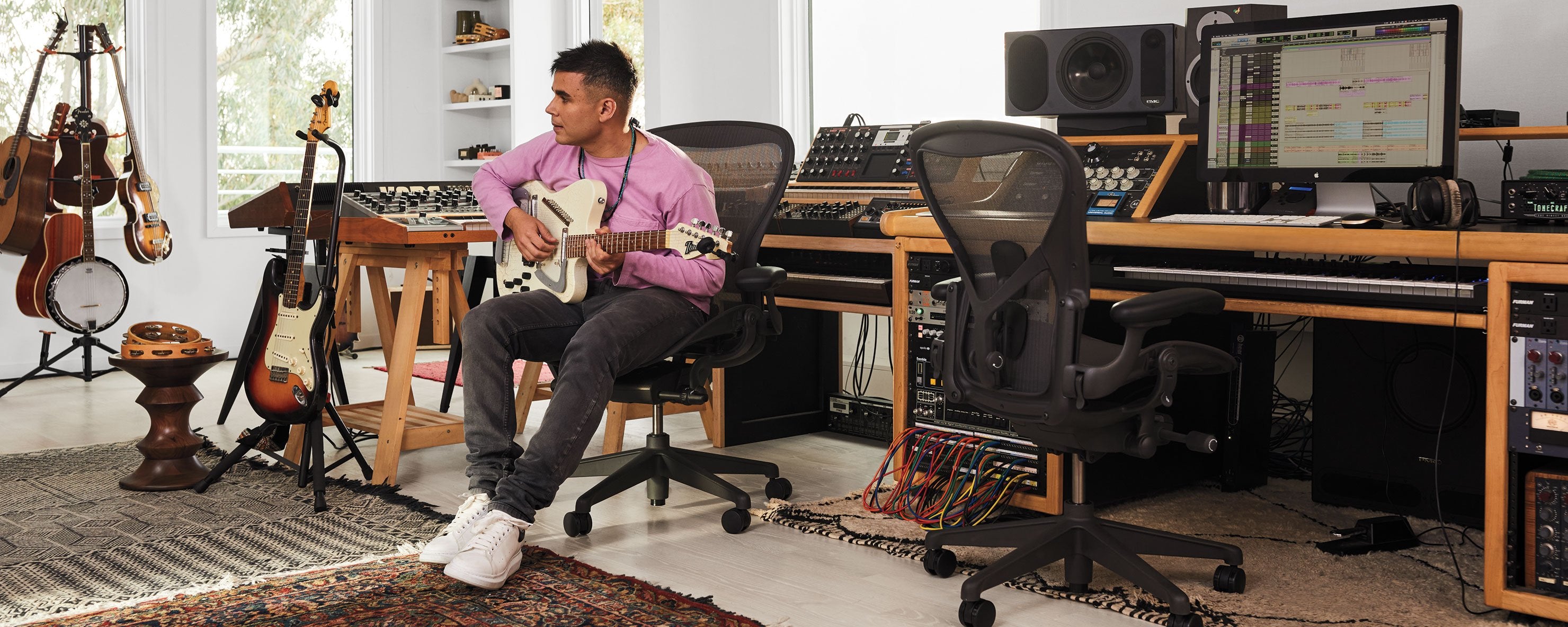 Photo of Rostam, muscian, producer and composer, in his music studio playing guitar sat in Aeron office chair in graphite