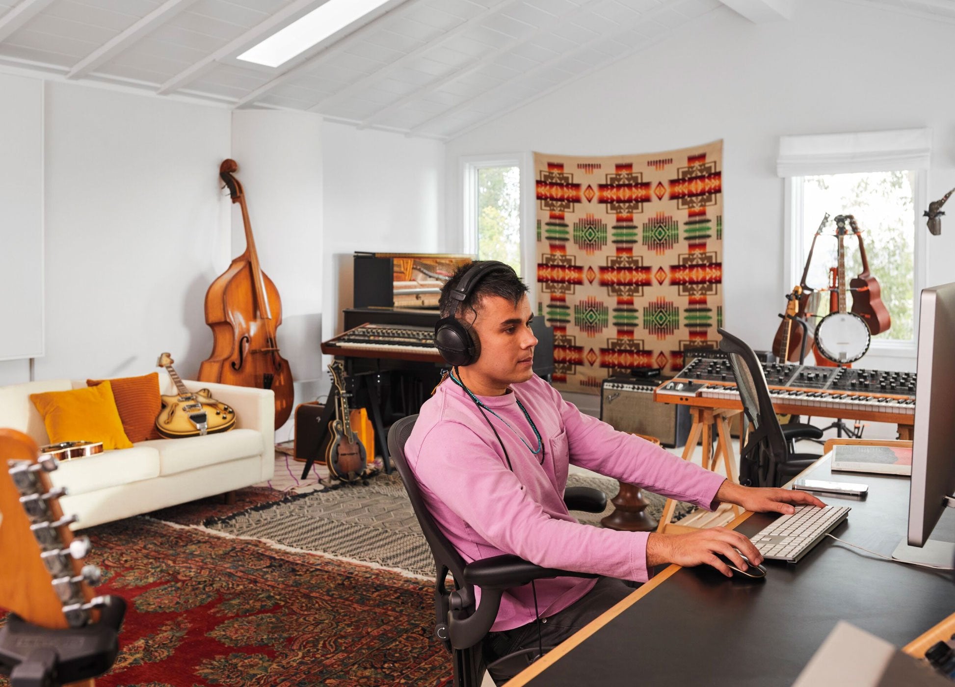 Photo of Rostam, muscian, producer and composer, sat in Aeron office chair in graphite at computer with headphones on with musical instruments in background